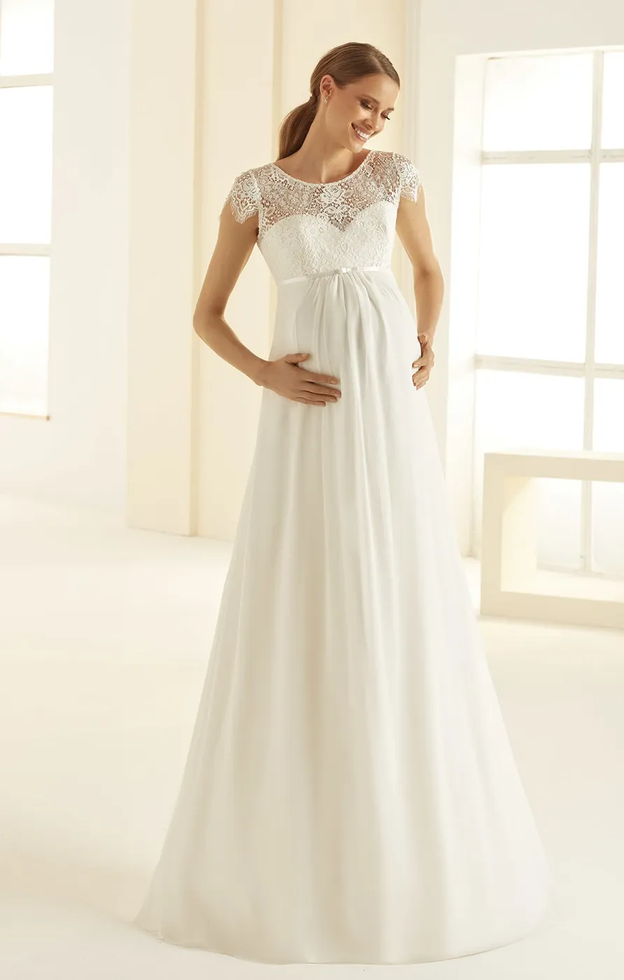 Schwangere Braut in fliessendem weissem Chiffon Brautkleid mit Spitzenoberteil und kurzen Ärmeln in heller romantischer Studio Kulisse