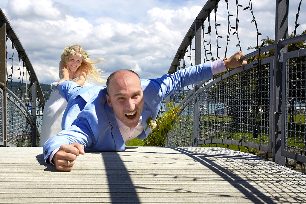 Zur Hochzeitsfotografie Eine Braut im weissen Brautkleid zieht den Bräutigam am Bein von deiner Brücke, blauer Himmel und fröhliche Stimmung