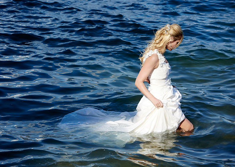 Braut watet im Brautkleid durch den See