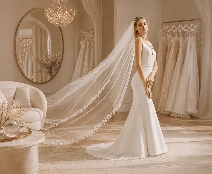 Braut im eleganten Brautkleid mit langem Schleier im hellen Showroom mit Brautkleidern in warmen beige Farbtönen